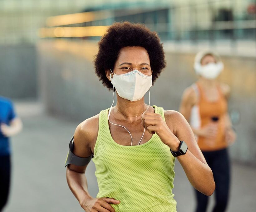 Mulher negra, com cerca de 30 anos, usando máscara e praticando corrida em local público.